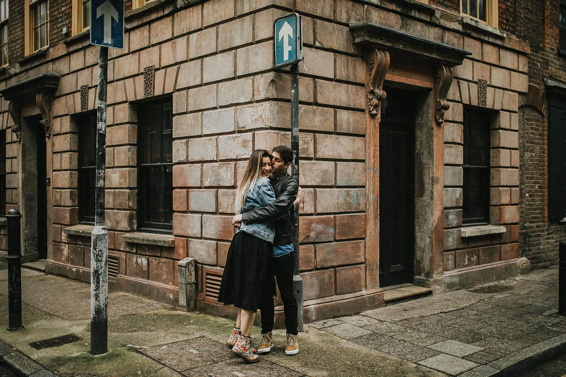 In an old city alley, a couple shares an embrace on the cobblestone sidewalk. The woman, in a black skirt and denim jacket, is smiling as she hugs the man, who is wearing a black leather jacket and jeans. Behind them, a vintage building with brown stone façade and barred windows adds a historical touch to the romantic scene. An arrow sign pointing upwards adds a symbolic element to the composition.