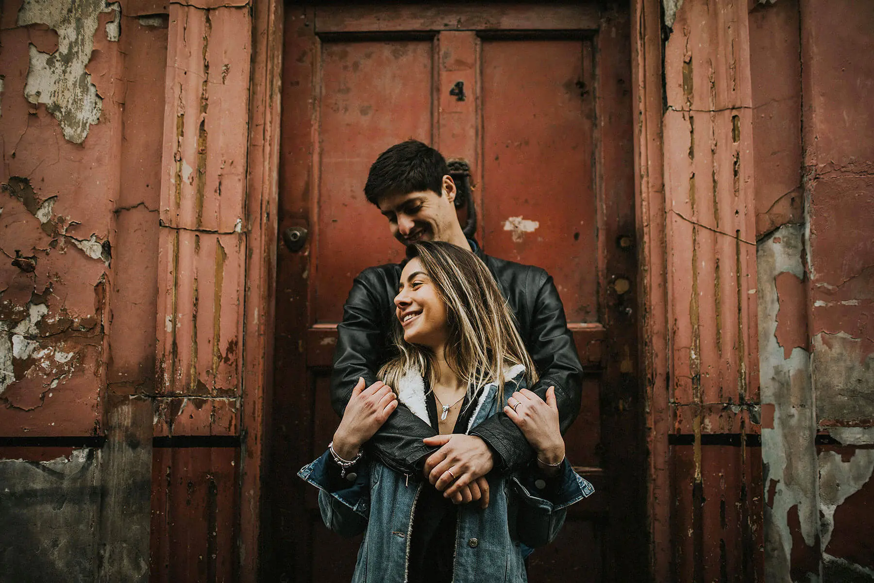 A couple stands in front of a weathered red door with peeling paint, sharing a moment of joy. The woman is in the foreground, laughing and looking to the side, while the man stands behind her, smiling down at her affectionately with his arms wrapped around her shoulders. Both are dressed casually in leather and denim jackets, adding a relaxed vibe to the intimate scene. 