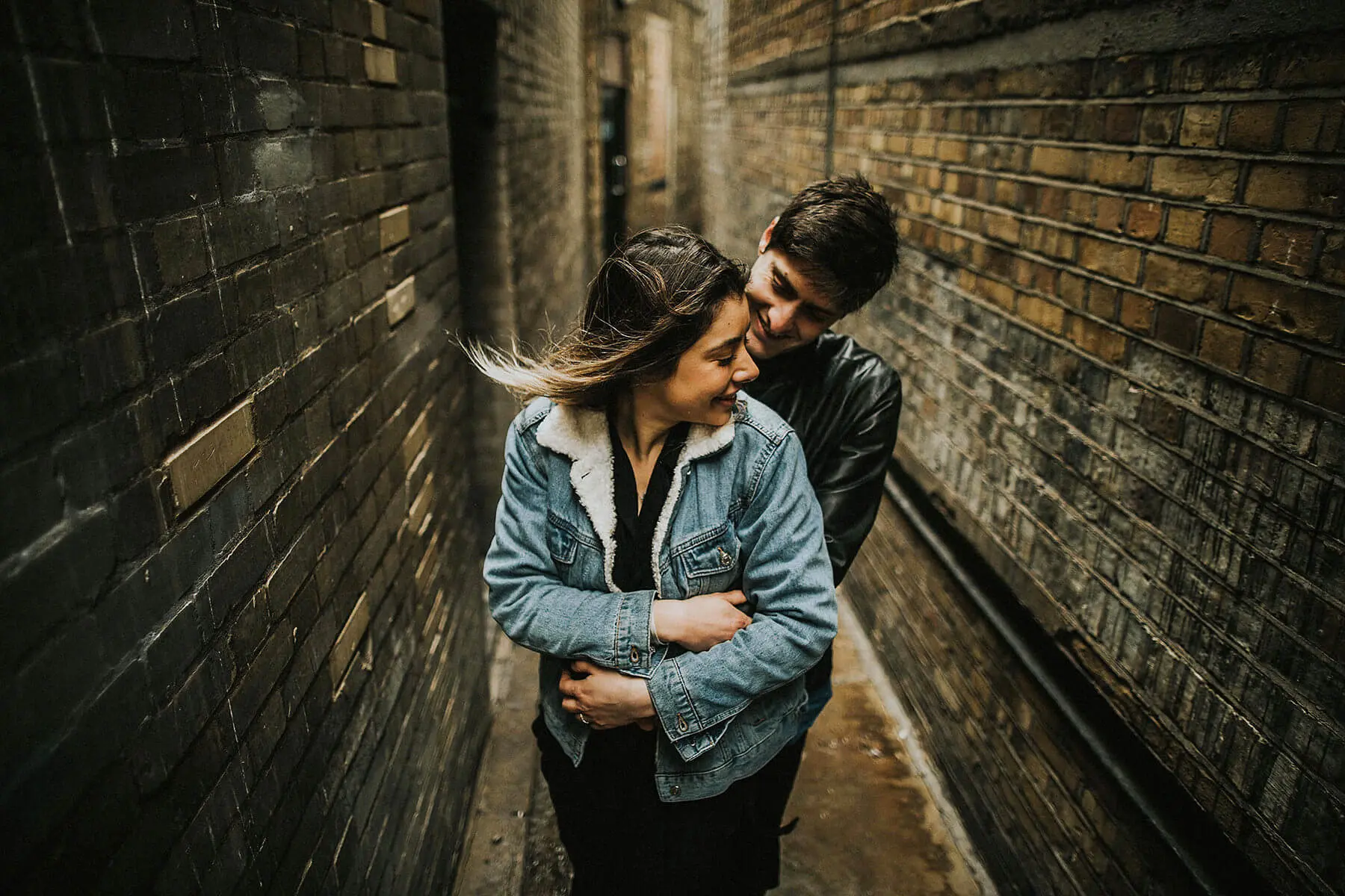 A smiling couple is embraced in a narrow alley with brick walls. The woman, wearing a denim jacket with a fur collar, looks content as the wind gently blows her hair. The man, in a black leather jacket, hugs her from behind, resting his chin on her shoulder with a joyful expression. The warmth between them contrasts with the urban, gritty setting.