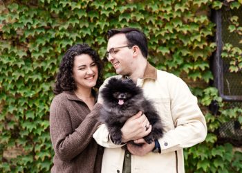 Couple holds Pomeranian while smiling in front of ivy wall during their Ravenswood neighborhood engagement session