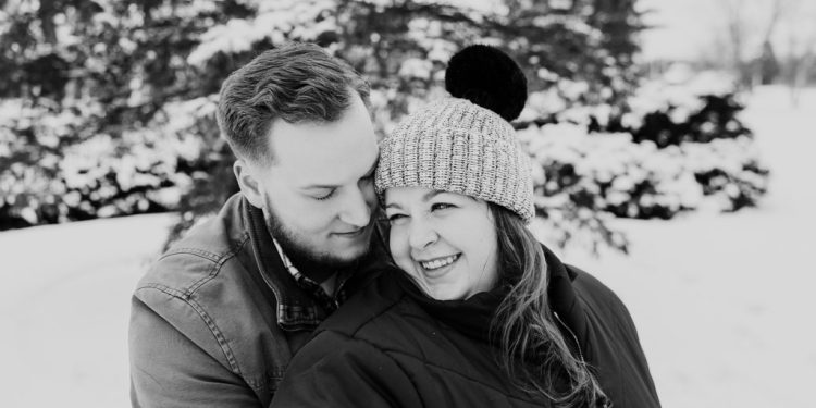 Candid photo of happy couple in coats and hats keeping each other warm during snowy winter engagement session