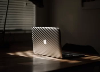 A MacBook Air is sitting on a table lit up by rays of sun through a window shade