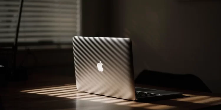 A MacBook Air is sitting on a table lit up by rays of sun through a window shade