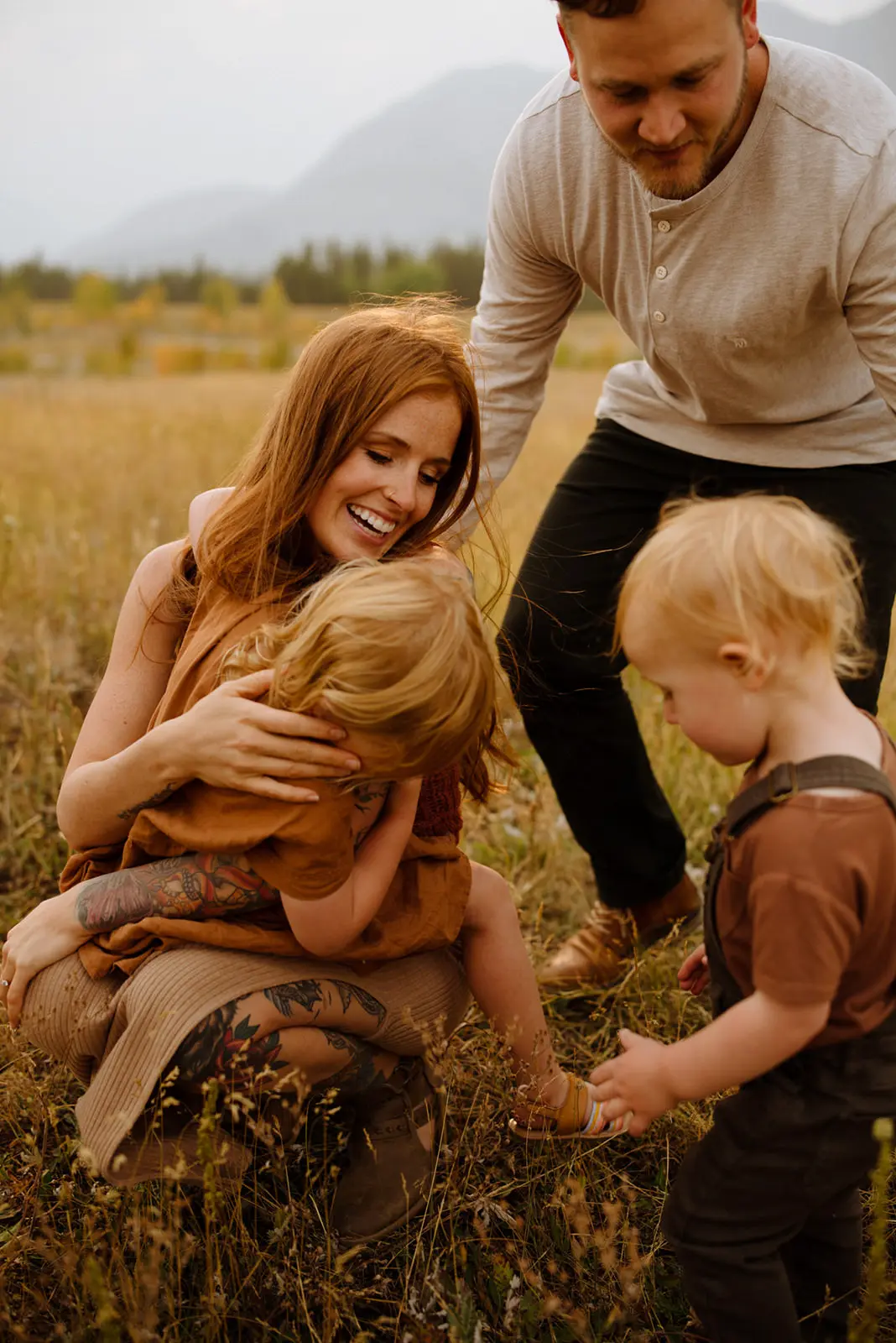 Joyful family moment in a golden field, with a smiling redhead mother embracing her laughing child, as a toddler reaches out to them and the father looks on, all set against a mountainous backdrop.