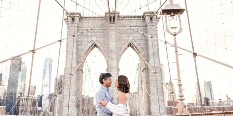 Creative Brooklyn Bridge engagement photo of couple standing on bridge with New York City skyline at sunset