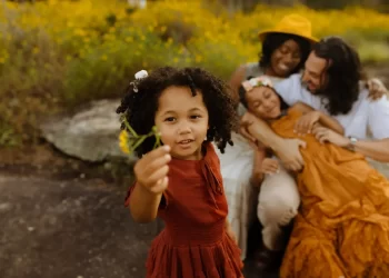 ZION Presets by Meridian enhance this family portrait of a young girl with curly hair holding a flower, with her family lovingly interacting in the background in a field of yellow flowers.