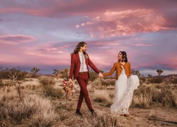 A couple walking hand-in-hand at sunset in Joshua Tree National Park, showcasing an all-inclusive wedding packages. The bride wears a white gown with a brown jacket, while the groom sports a burgundy suit, holding a bouquet of flowers.